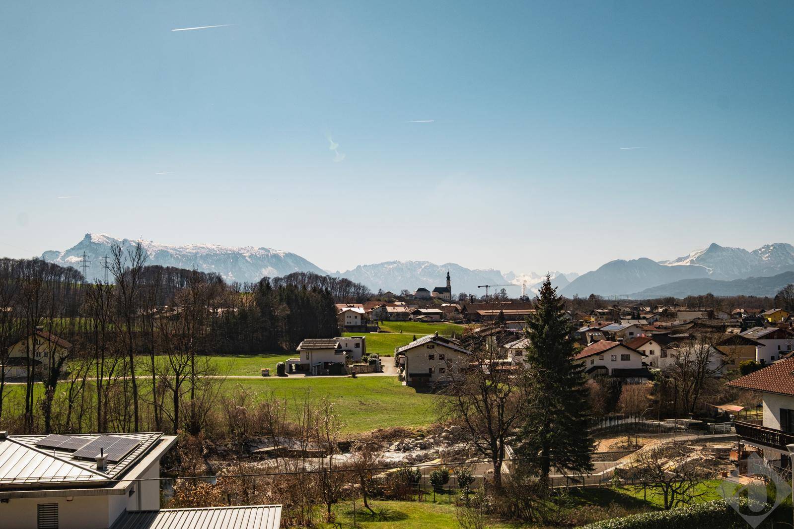 Panoramablick über eine ländliche Umgebung mit Wohnhäusern, grünen Feldern und majestätischen Bergen am Horizont.