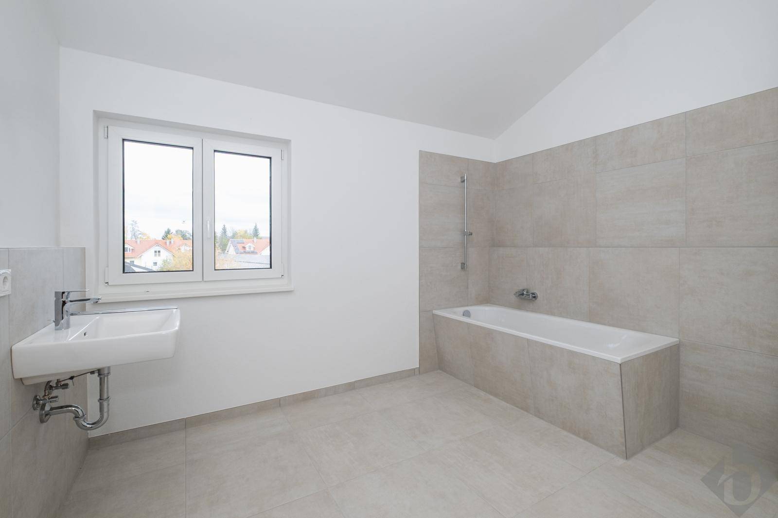 Modernes Badezimmer mit Badewanne, Waschbecken, Fenster und hellgrauen Fliesen an Boden und Wänden.