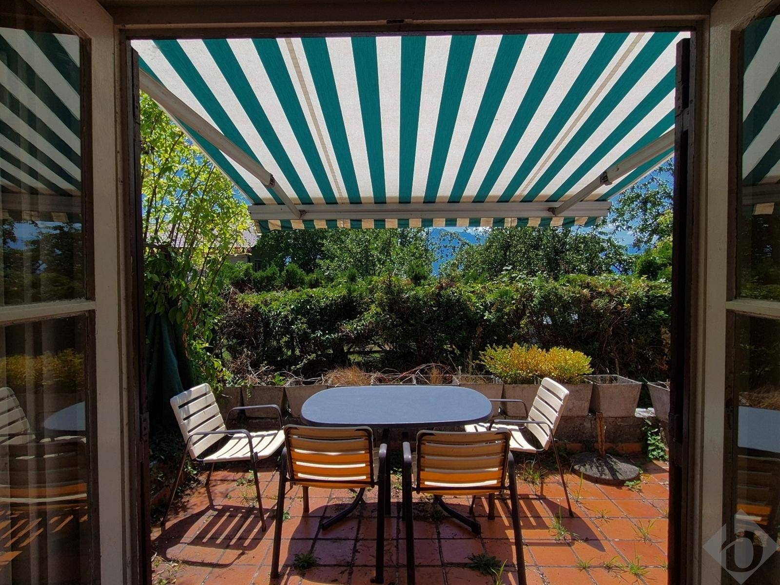 Sonniger Terrassenbereich mit Esstisch, Stühlen und einer gestreiften Markise, umgeben von viel Grün.
