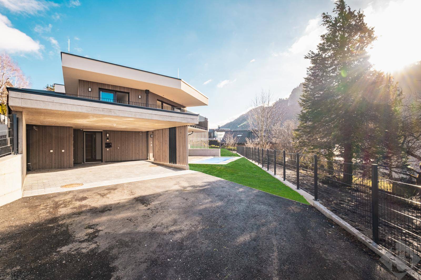 Moderne Architektur mit Carport und Zufahrt, umgeben von grüner Landschaft und Bergen.
