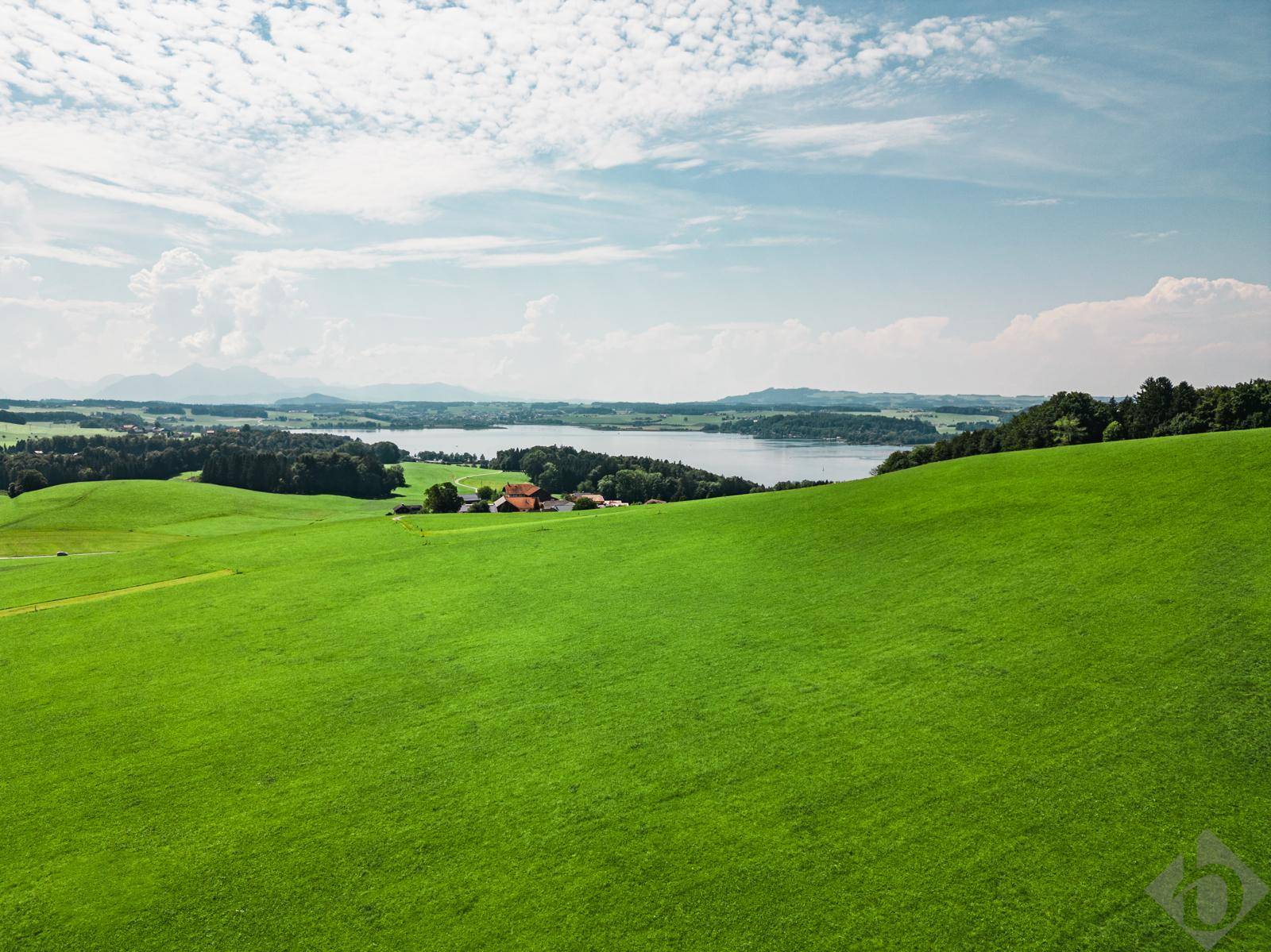 Weitläufige grüne Hügellandschaft mit einem See und Bergen am Horizont unter blauem Himmel.