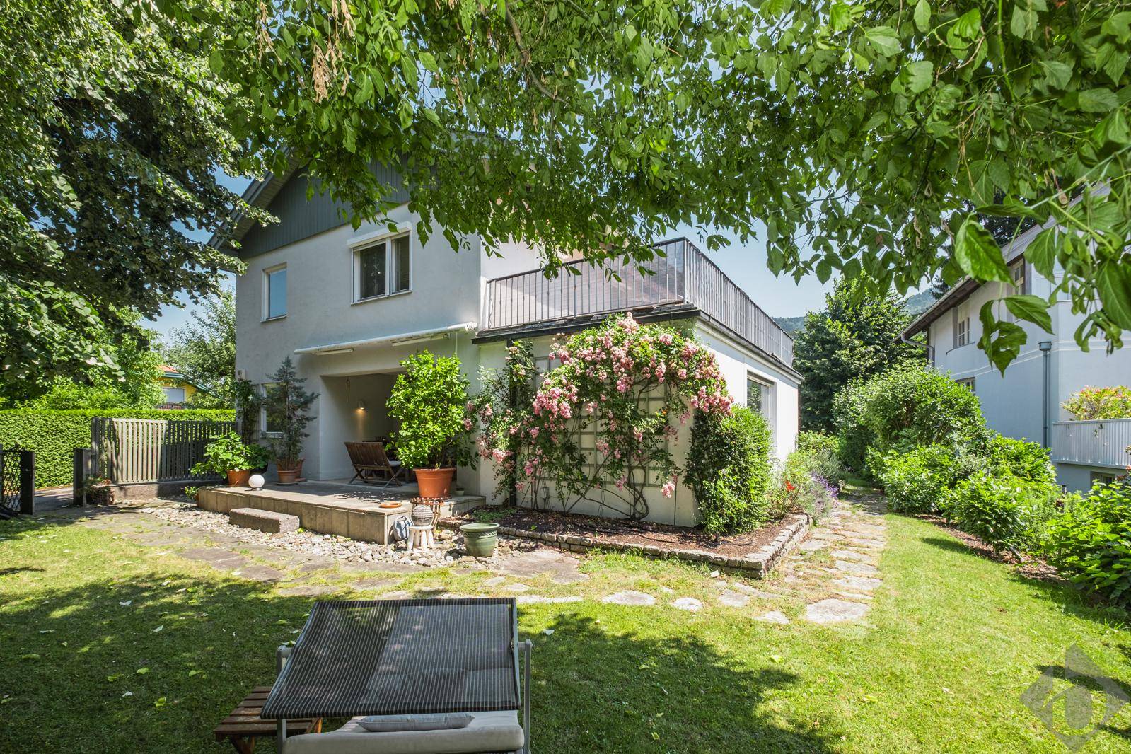 Ruhiger Gartenbereich mit überdachter Terrasse, blühenden Pflanzen und viel Grün.