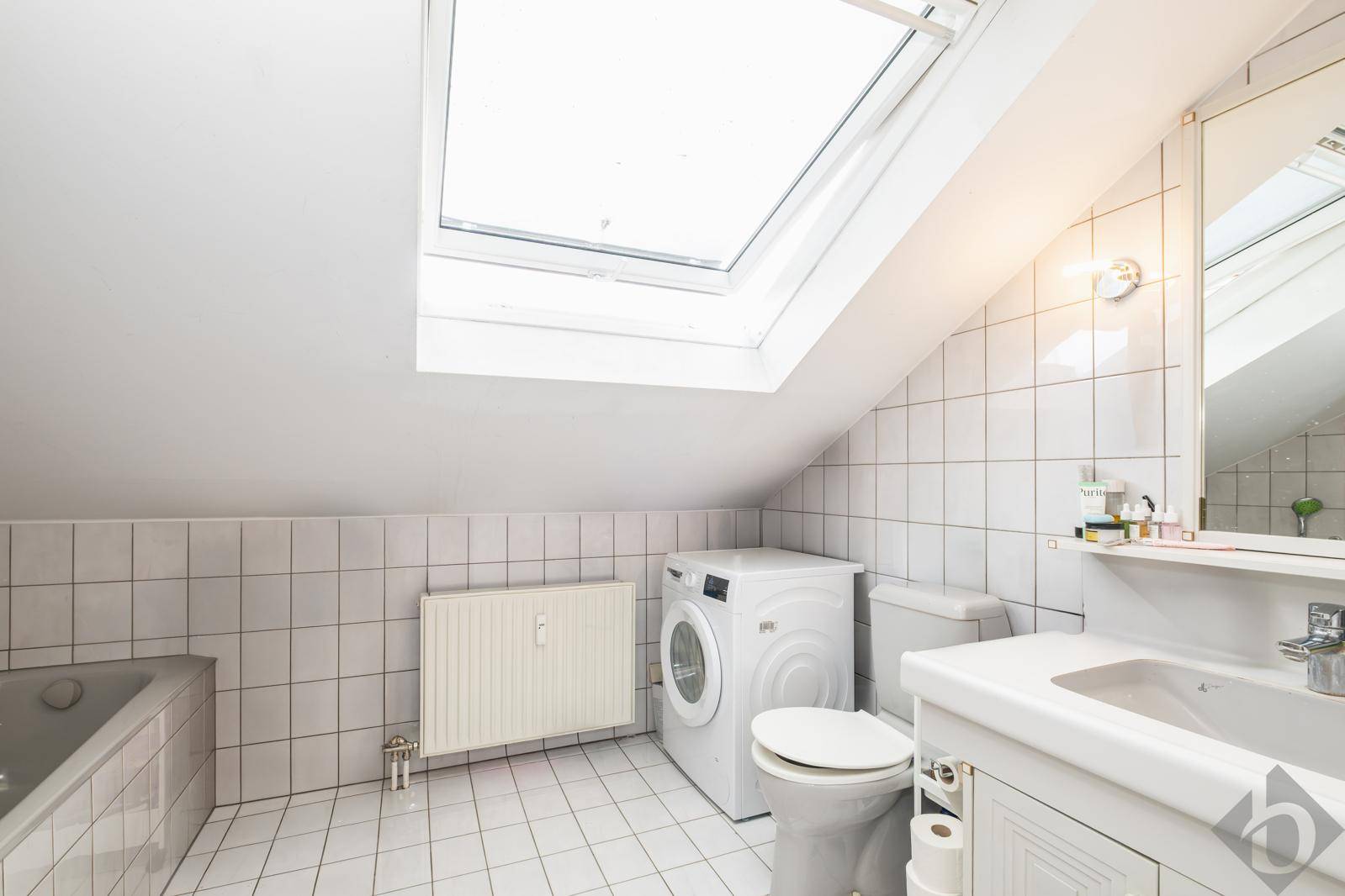 Badezimmer im Dachgeschoss mit weißen Fliesen, Badewanne, Waschmaschine und großem Dachfenster.