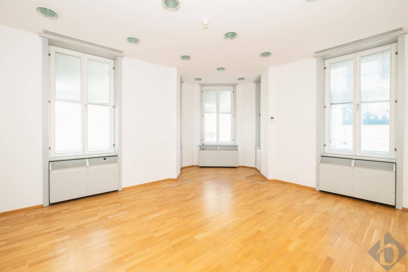 Leerer Büroraum mit hellem Holzfußboden und mehreren Fenstern, die viel Tageslicht hereinlassen.