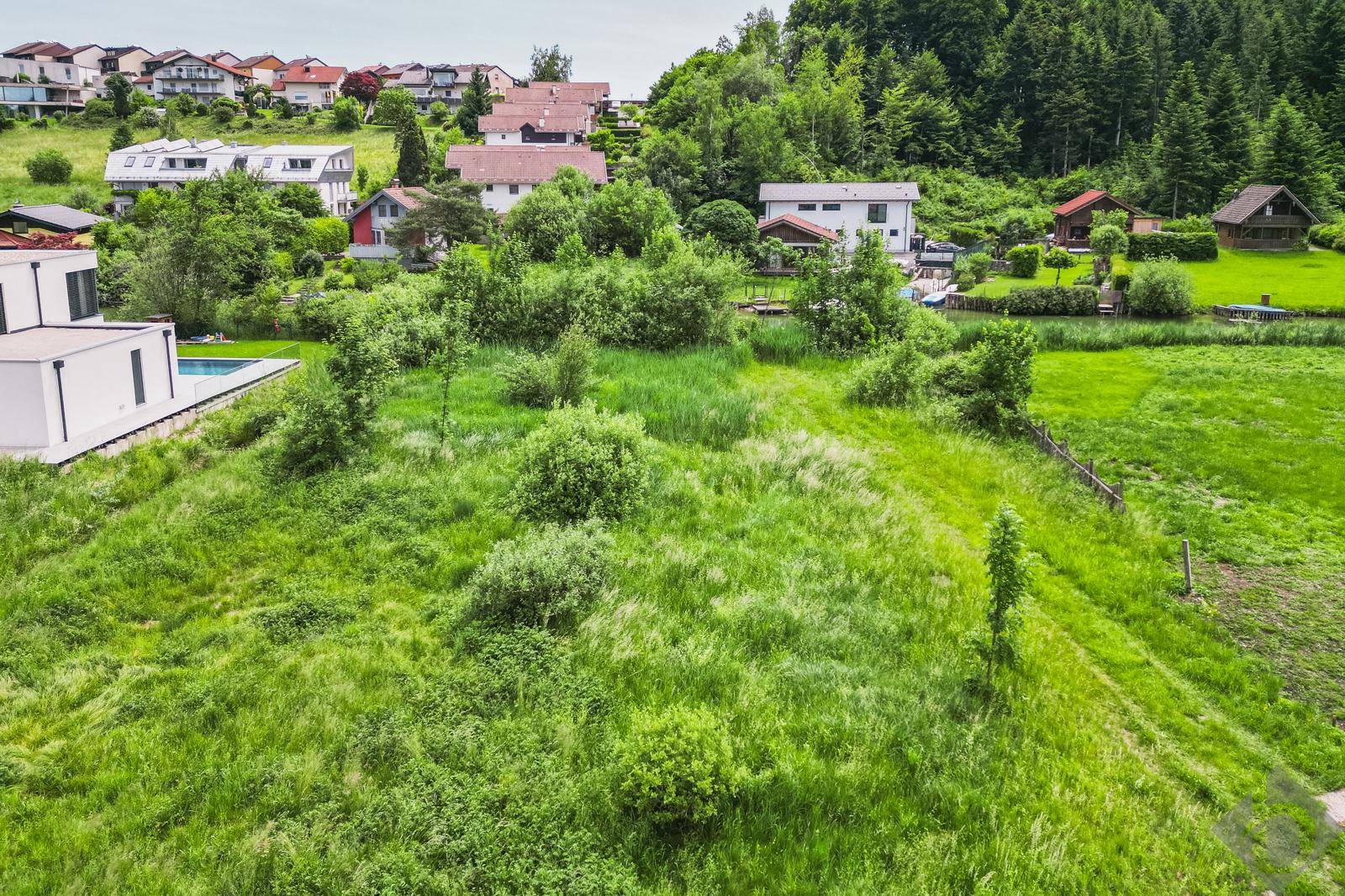 Weitläufige grüne Wiese mit Blick auf eine Siedlung und bewaldete Hügel unter hellem Himmel.