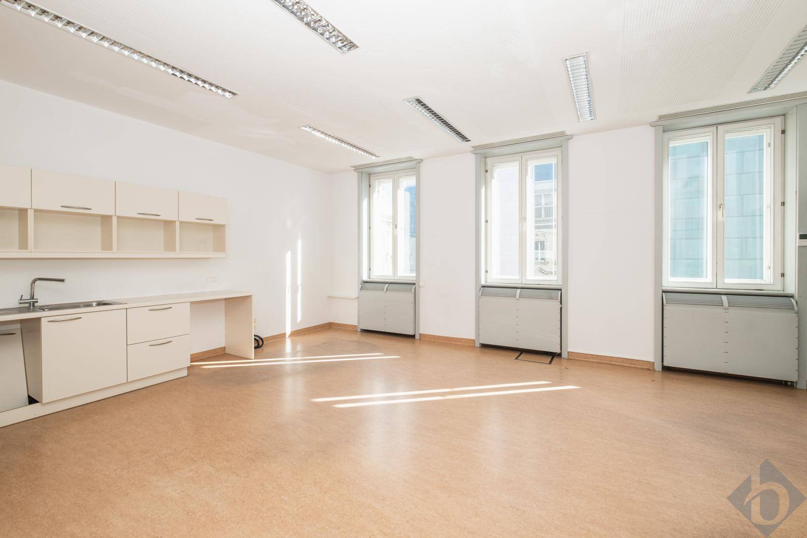 Heller Büroraum mit großen Fenstern, einer kleinen Küchenzeile und Linoleumboden, ideal für gewerbliche Nutzung.