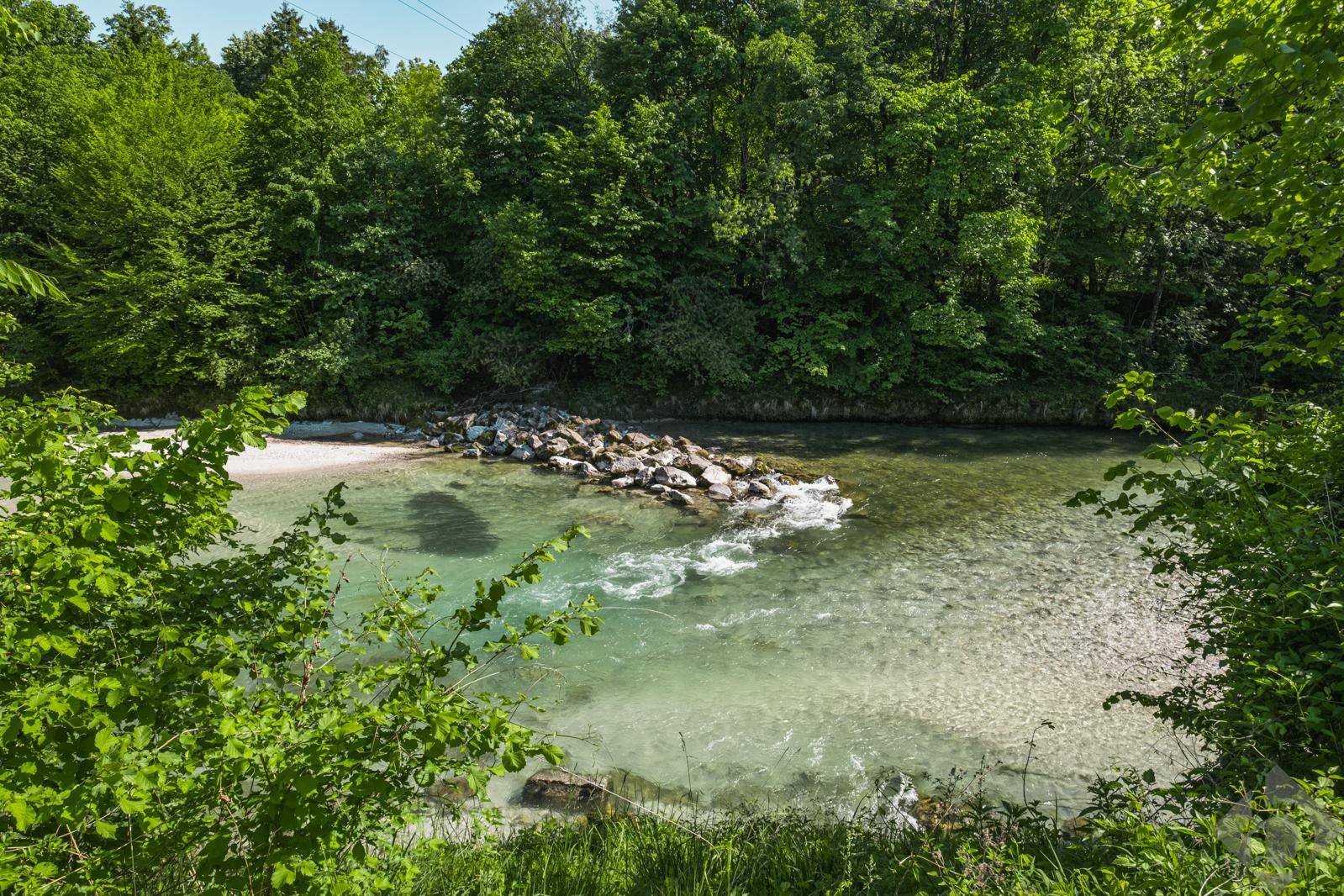 Klarer Fluss mit Felsen im Wasser, umgeben von dichtem Grün und Bäumen.