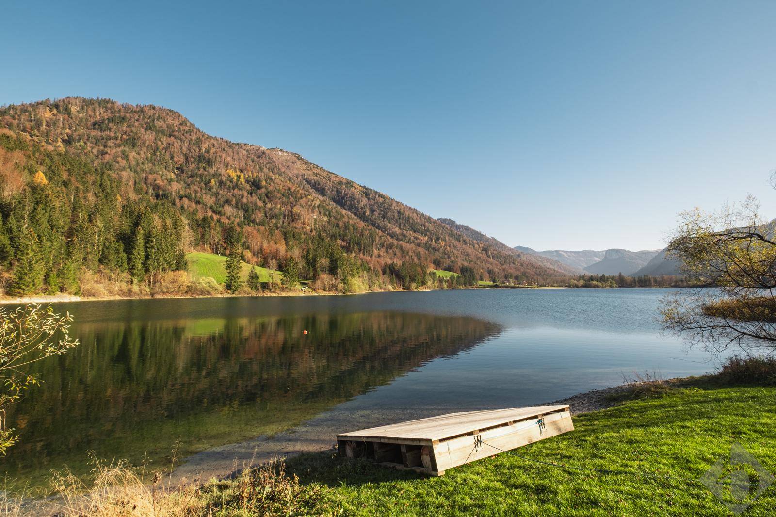 Ruhiger See mit einem kleinen Holzsteg am Ufer, umgeben von bewaldeten Bergen und klarem Wasser.