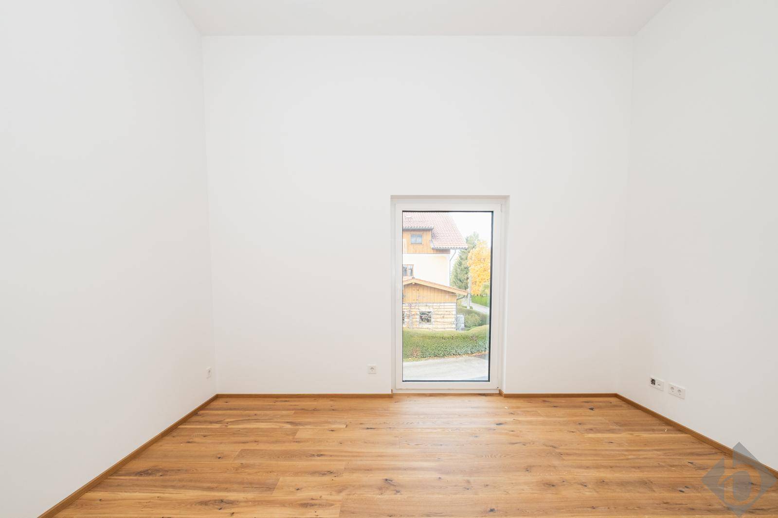 Heller, leerer Raum mit Holzboden, weißen Wänden und einer Glastür, die viel Tageslicht hereinlässt.