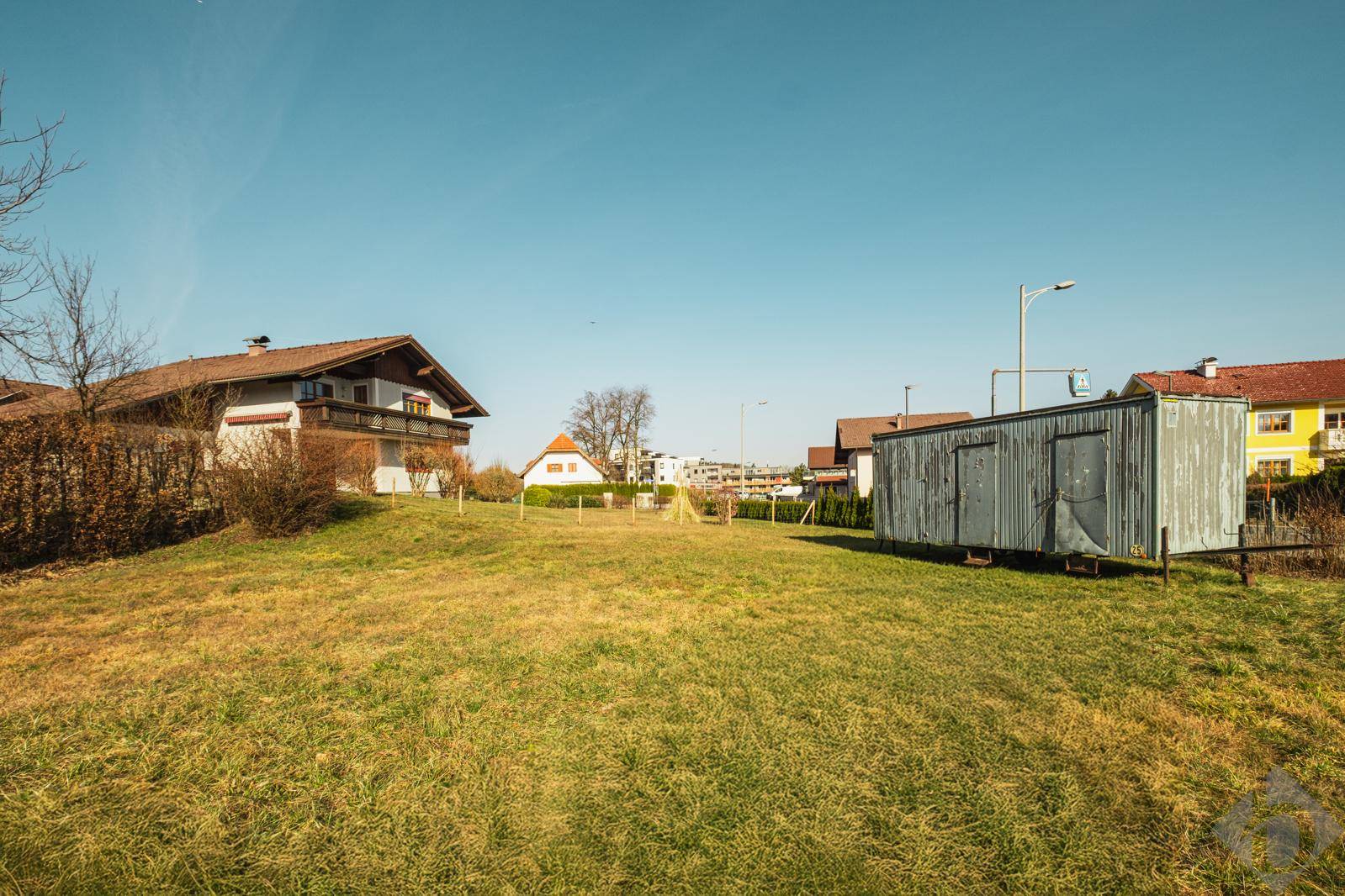 Blick über das grüne Grundstück mit einem Schuppen und umliegenden Wohnhäusern unter blauem Himmel.
