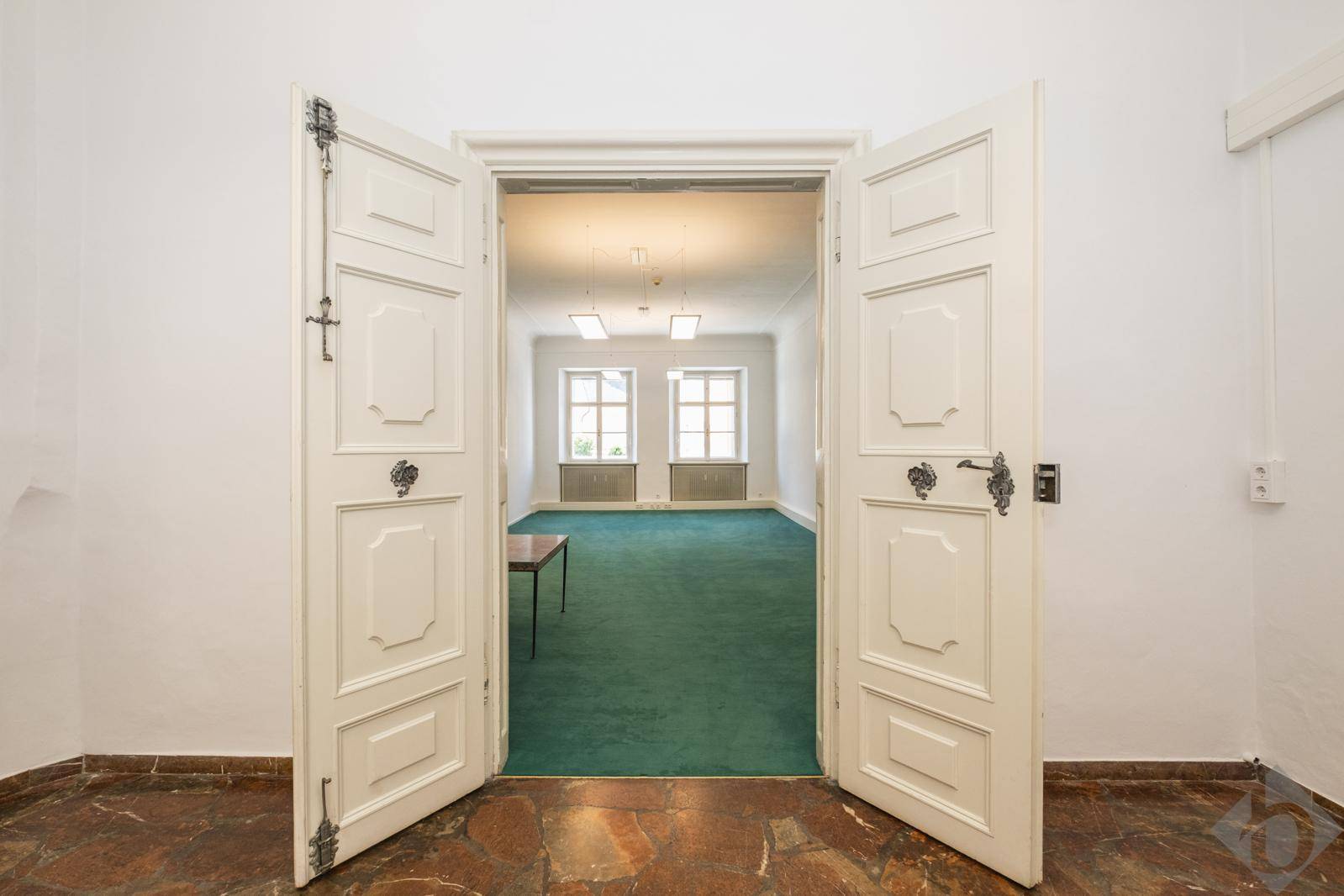 Blick durch eine offene Doppeltür in einen geräumigen Büroraum mit grünem Teppichboden und zwei Fenstern.
