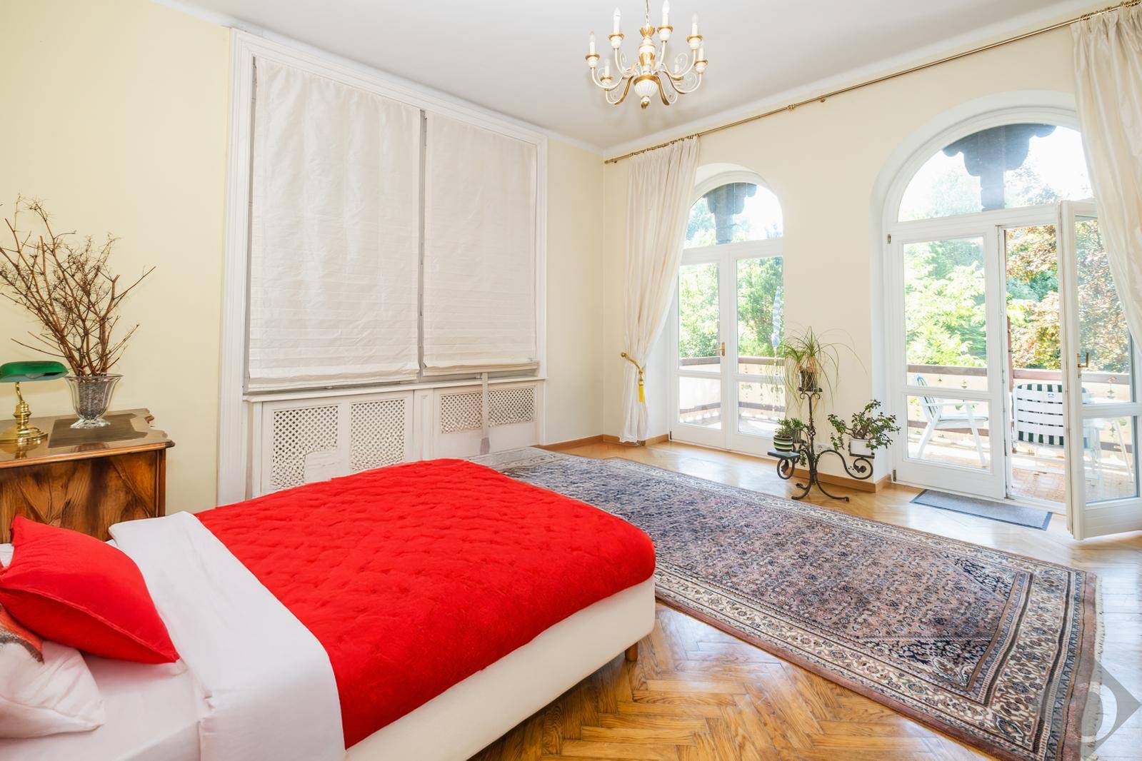 Helles Schlafzimmer mit roter Bettdecke, Fischgrätparkett und Zugang zu einem charmanten Balkon.