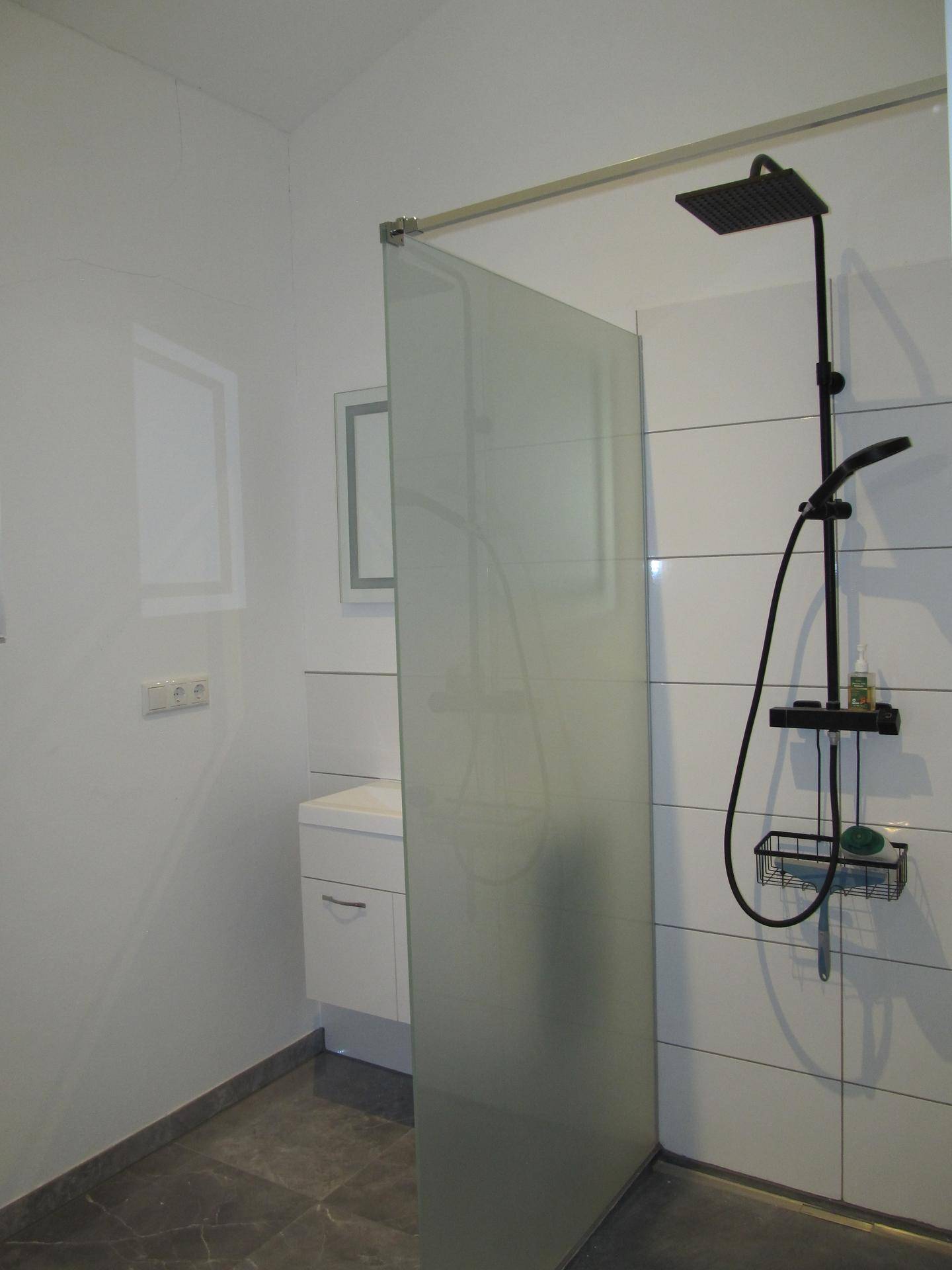 Modernes Badezimmer mit begehbarer Dusche, Regendusche und Waschtisch im Hintergrund.