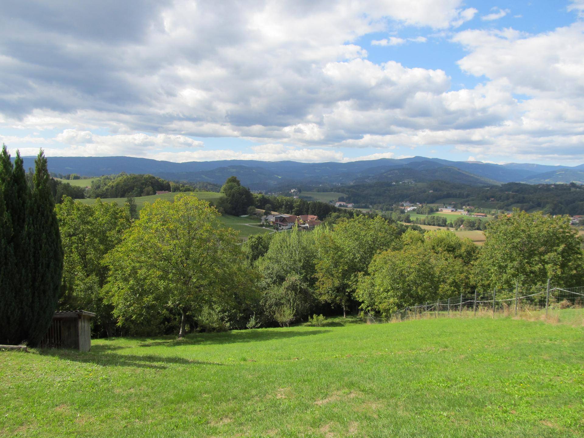 Panoramablick über eine weite grüne Landschaft mit Dörfern und Bergen am Horizont unter einem dramatischen Himmel.