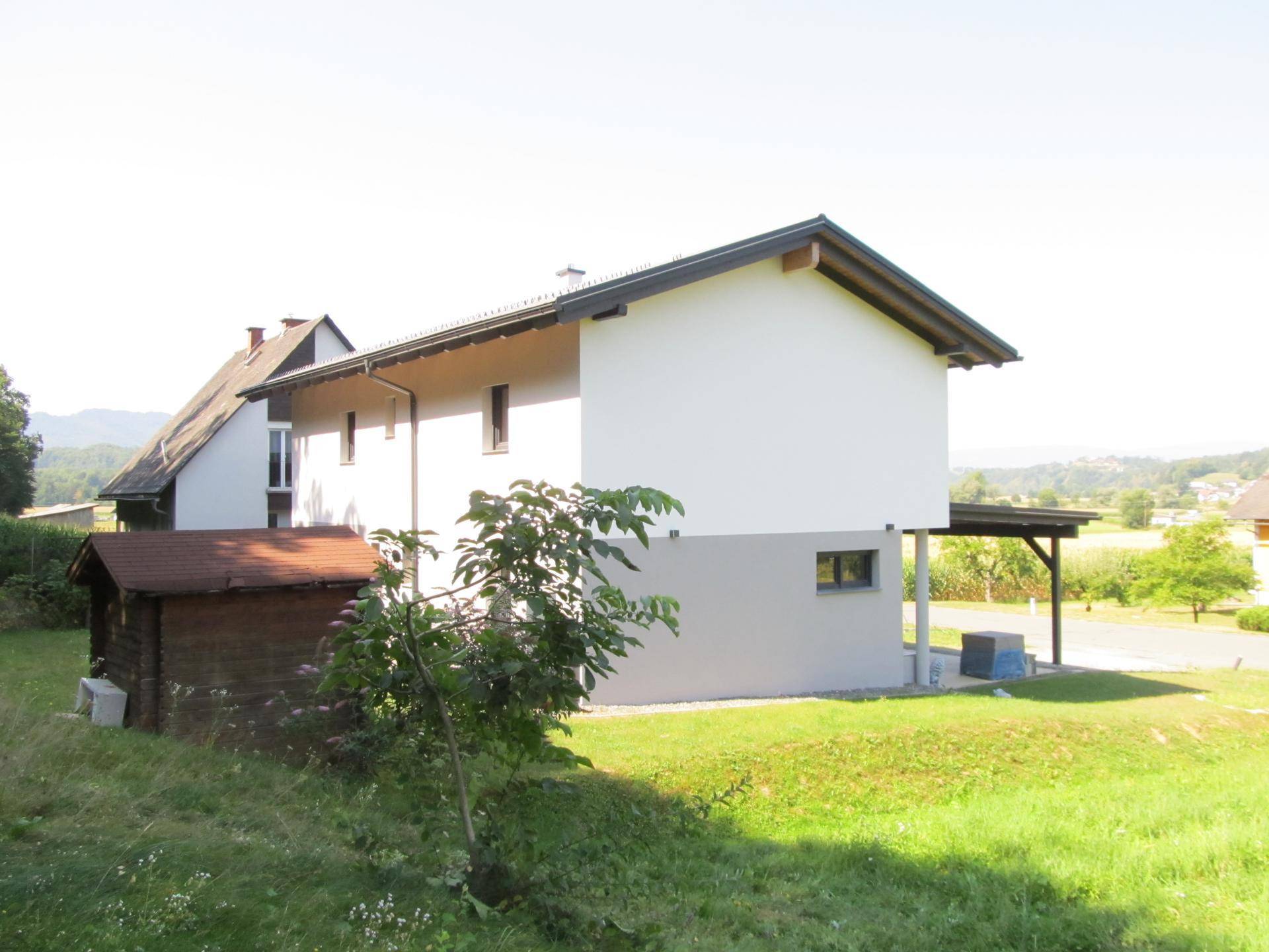 Seitenansicht des Hauses mit weißer und grauer Fassade sowie einem kleinen Holzschuppen.