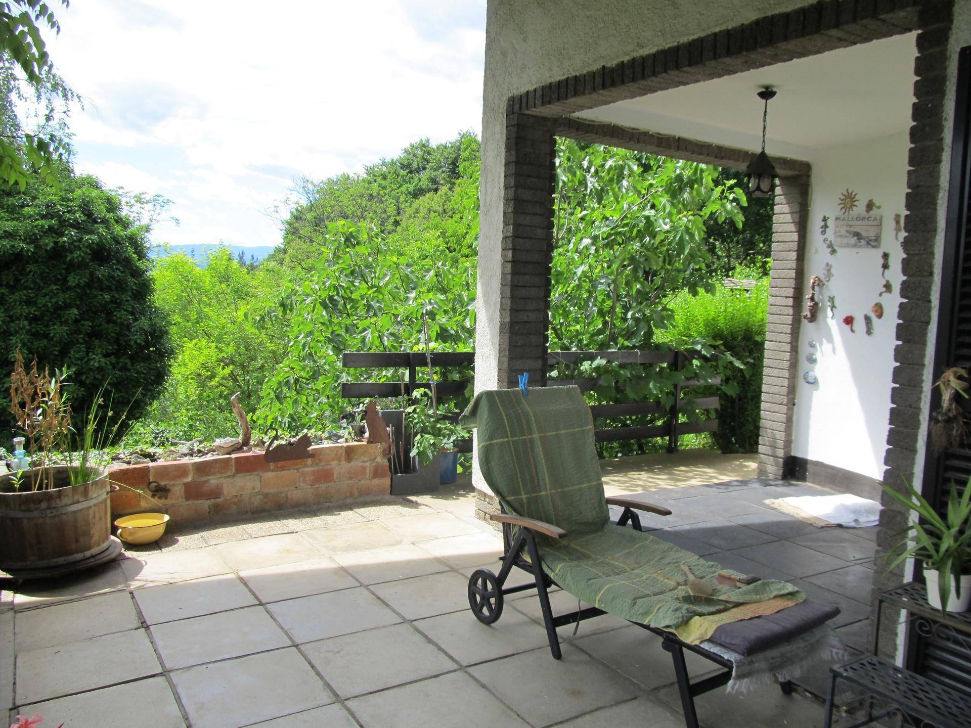 Überdachte Terrasse mit Fliesenboden, Liegestuhl und Blick auf die grüne Umgebung.