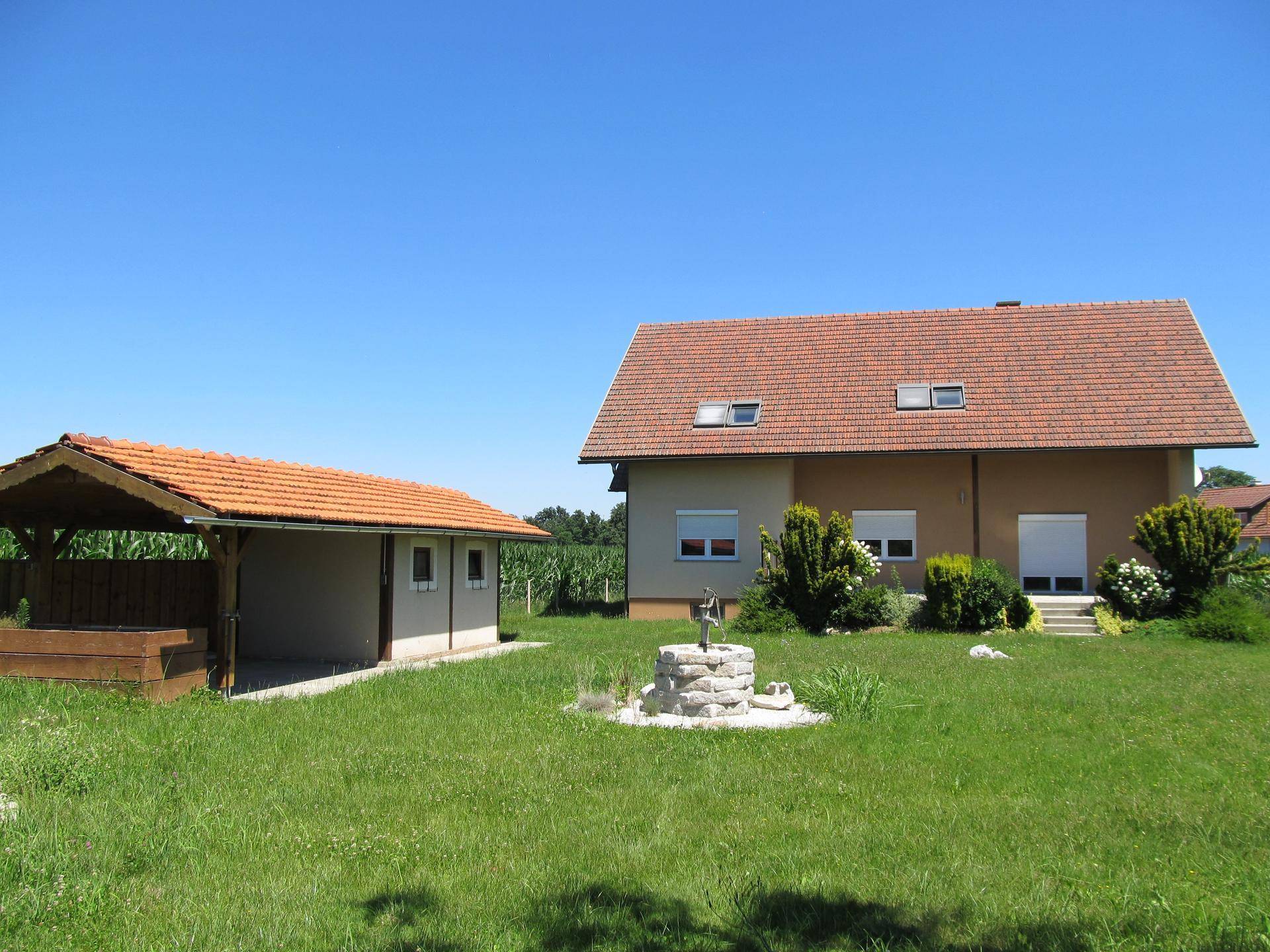 Gartenbereich mit Wohnhaus, Carport und Zierbrunnen, umgeben von grünen Feldern unter blauem Himmel.