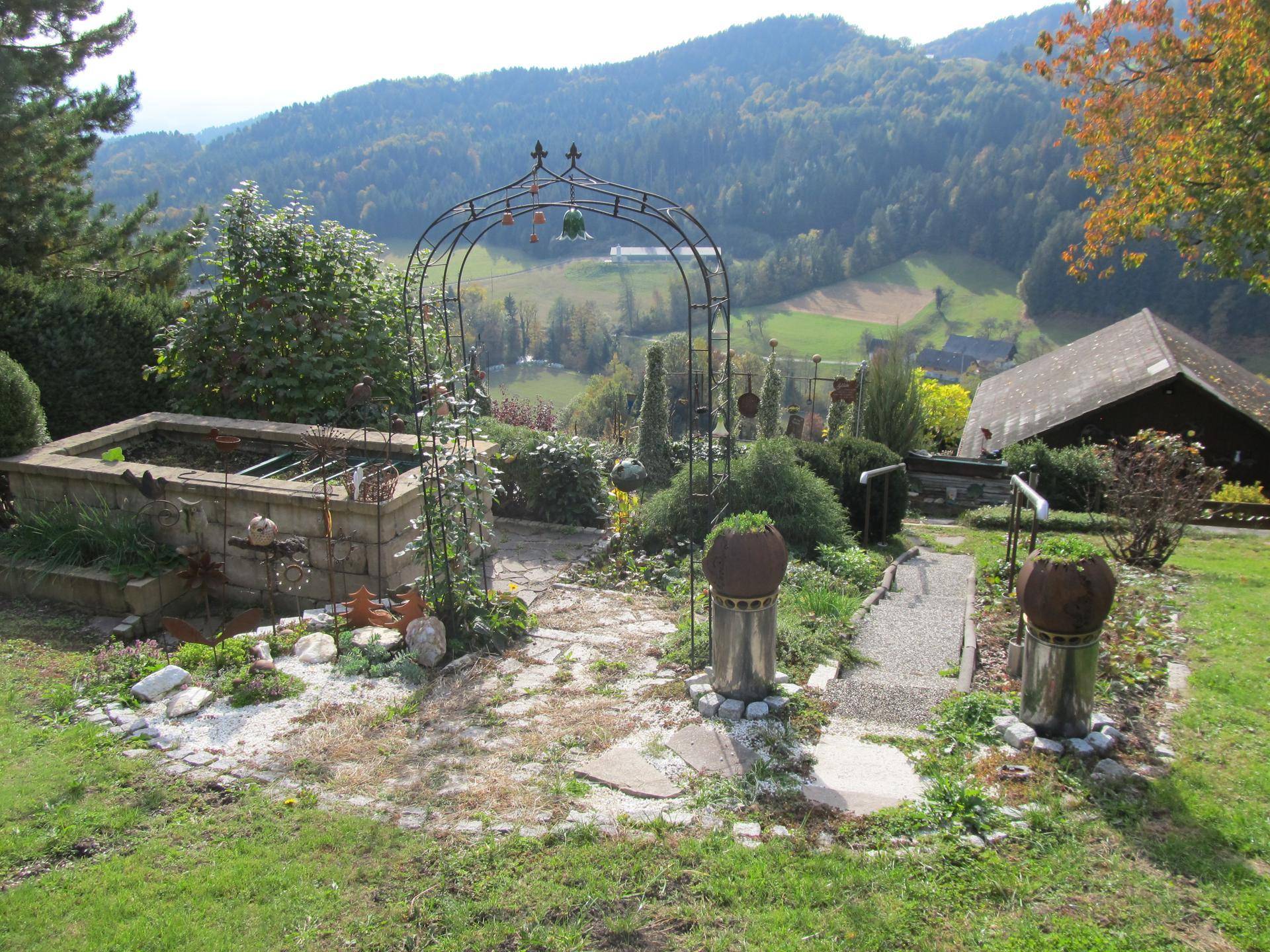 Terrassierter Garten mit Zierbrunnen, Metallbogen und weitem Blick über die Landschaft.
