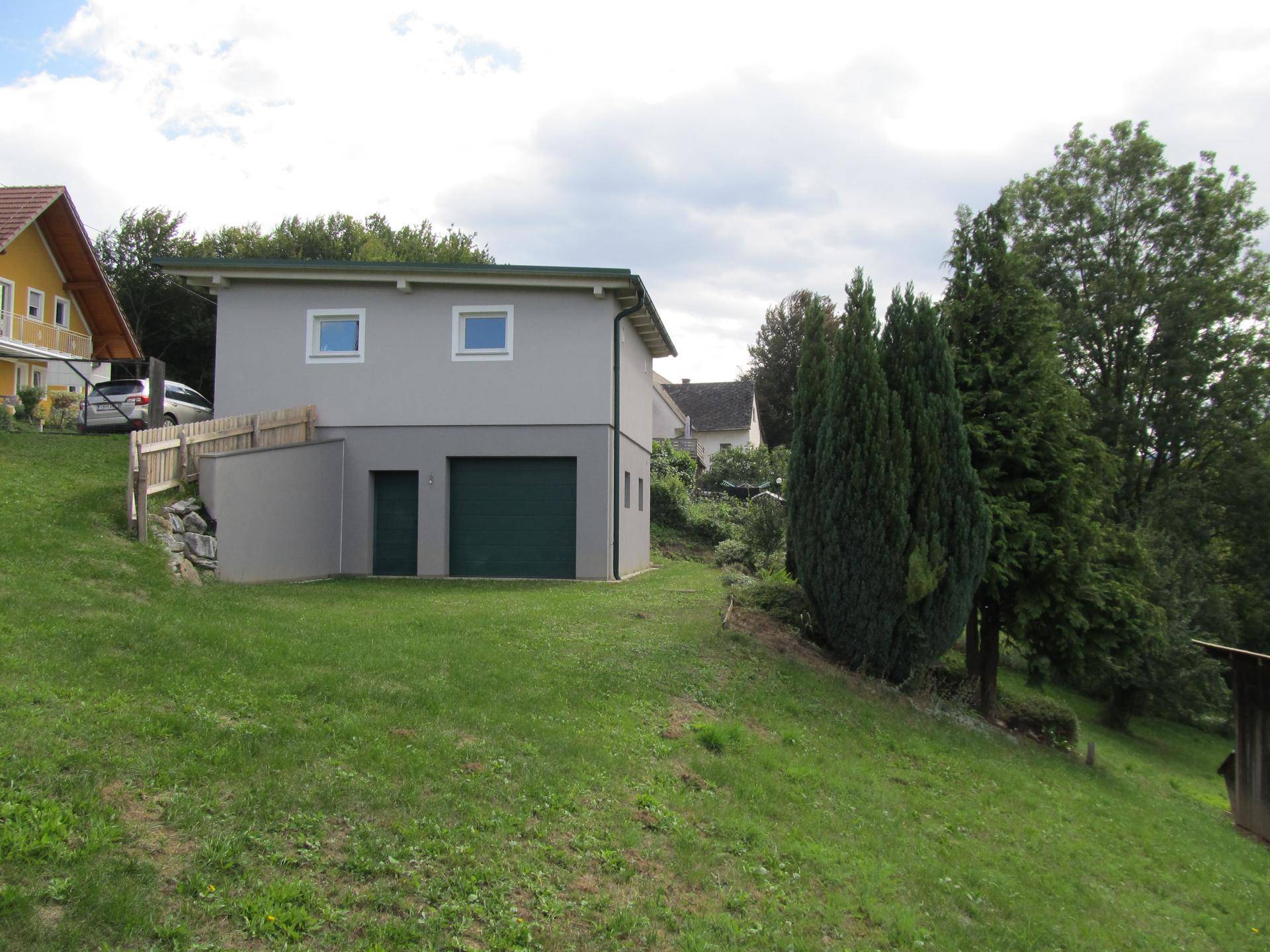 Ansicht des grauen Gebäudes mit Garage und kleiner Tür, umgeben von einem gepflegten Rasen und Bäumen.
