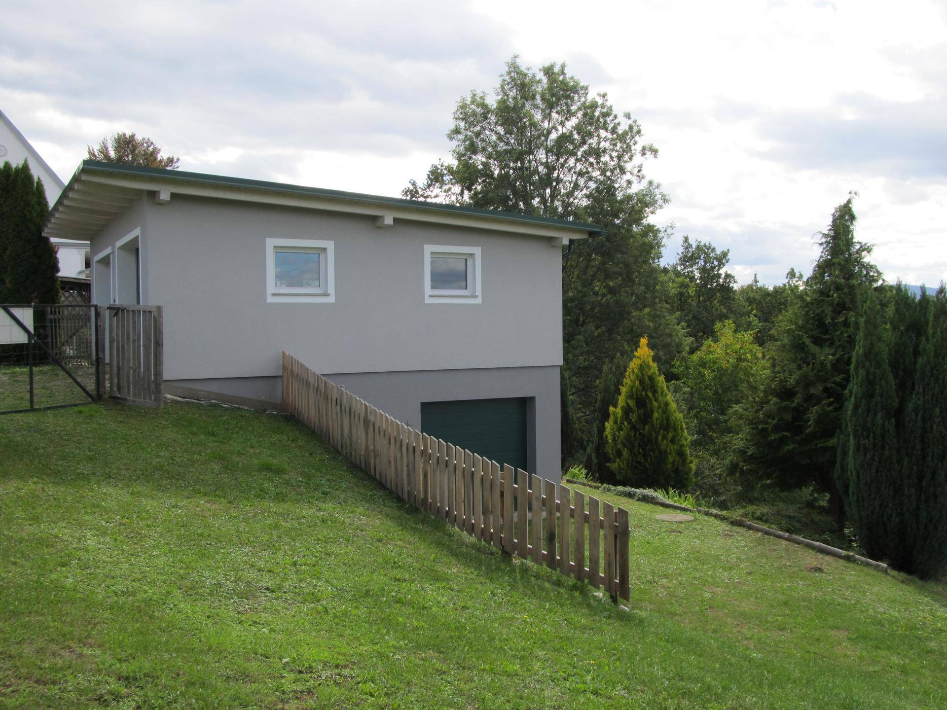 Seitenansicht des grauen Gebäudes mit zwei Fenstern und einer grünen Garage, eingebettet in eine grüne Landschaft.