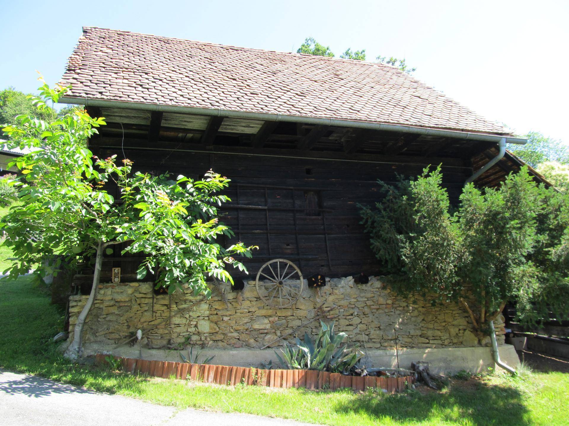 Altes Holzgebäude mit Ziegeldach und einer Steinmauer, umgeben von Bäumen und Sträuchern.