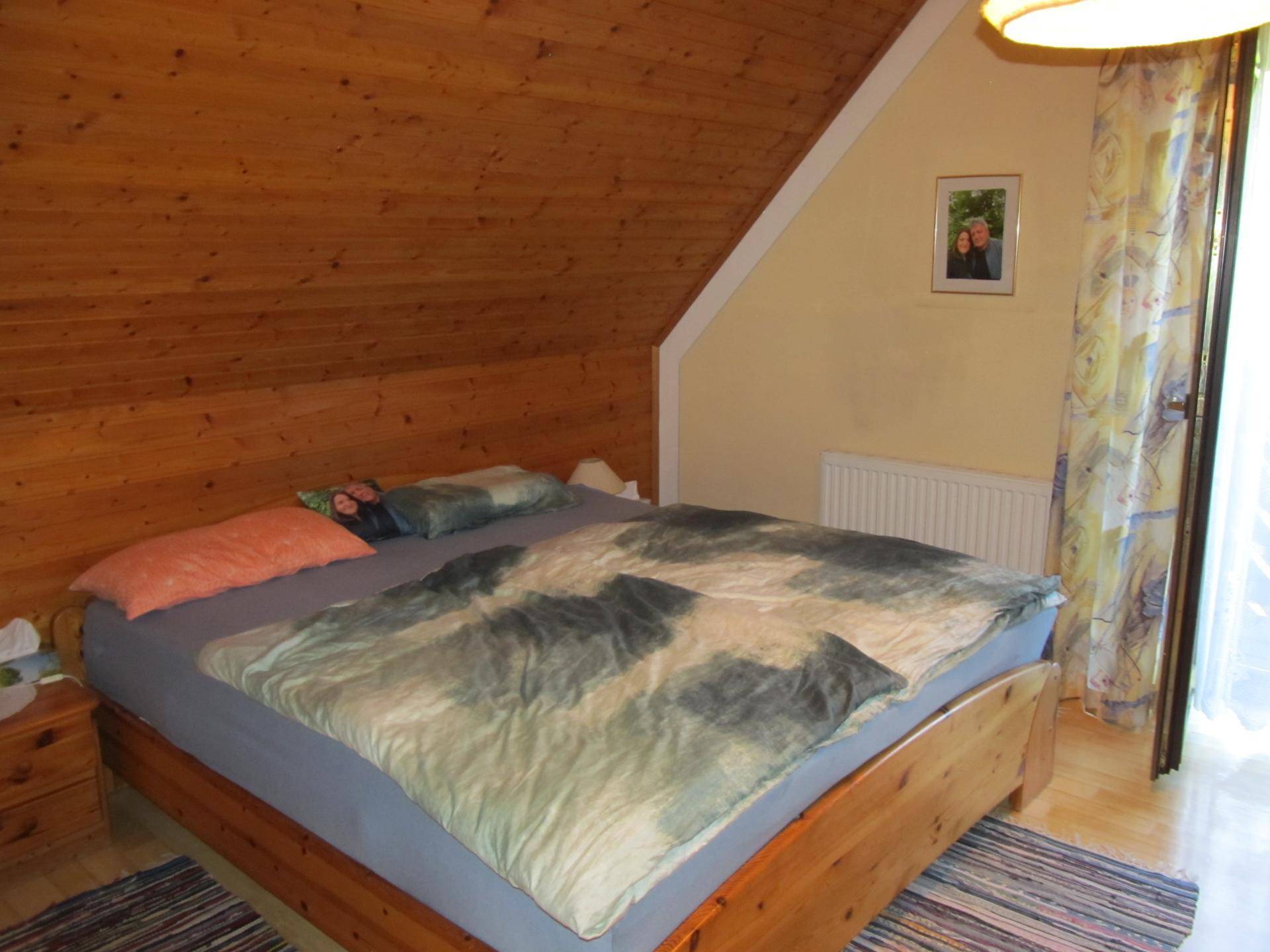 Gemütliches Schlafzimmer mit großem Holzbett, Holzdeckenverkleidung und Fenster mit Vorhängen.