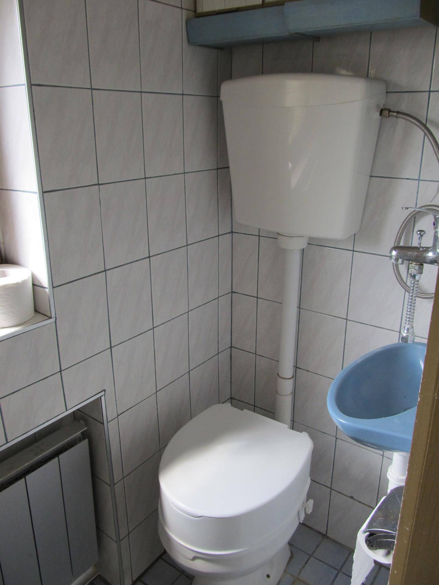 Kleines WC mit weißen Fliesen, erhöhtem WC und einem kleinen blauen Waschbecken.