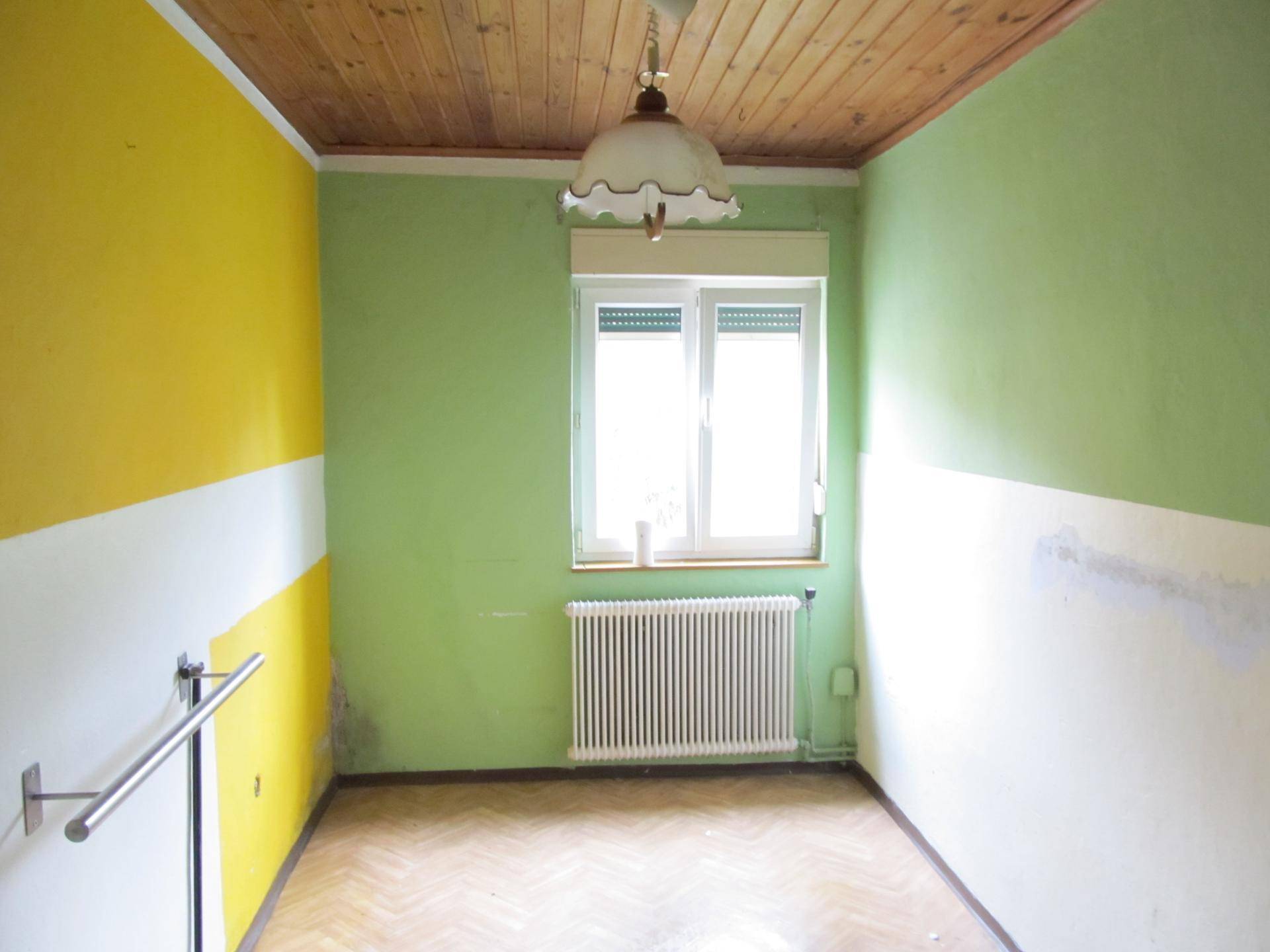 Helles Zimmer mit farbigen Wänden, Holzdecke, Parkettboden und einem großen Fenster mit Heizkörper.
