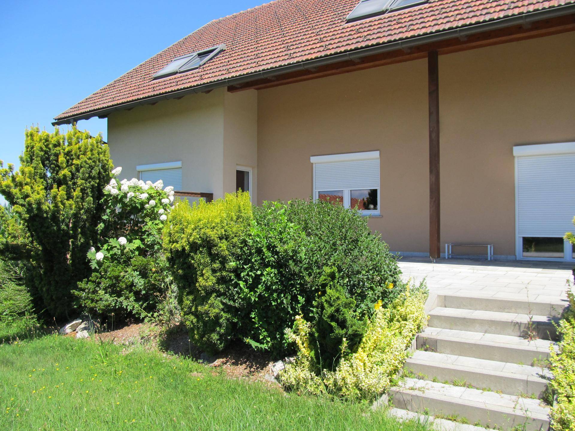 Seitliche Ansicht des Hauses mit gepflasterter Terrasse und Treppe zum grünen Gartenbereich.
