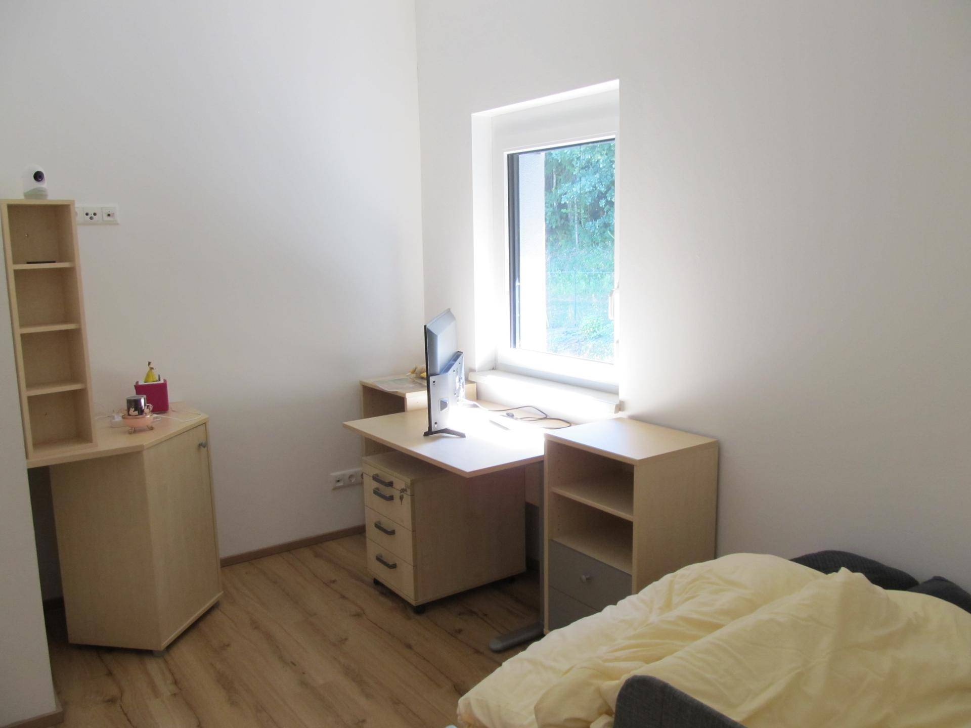 Schlafzimmer mit Schreibtisch, Regal und Fenster mit Blick ins Grüne.