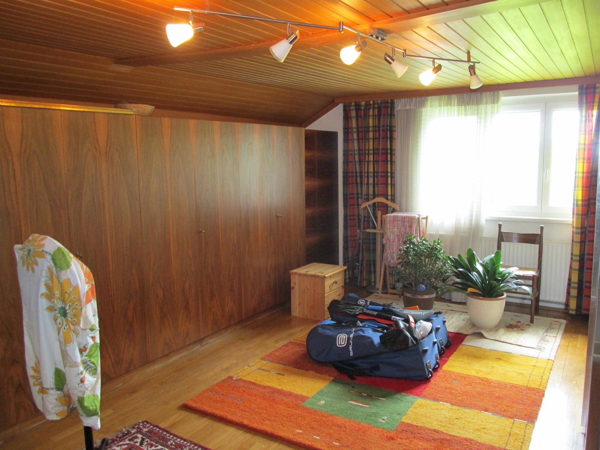 Zimmer mit Holzwänden und Holzdecke, Teppichboden und einem Fenster mit bunten Vorhängen.