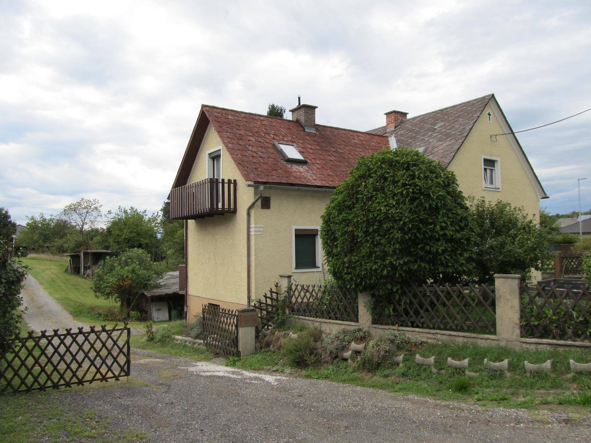 Rückansicht eines gelben Hauses mit rotem Dach, einem Balkon und einem Gartentor in ländlicher Umgebung.