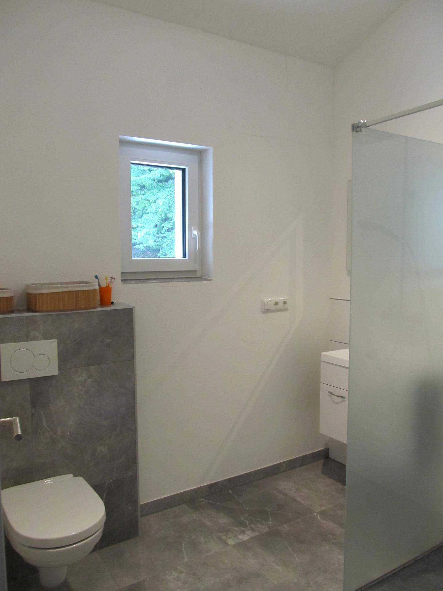 Helles Badezimmer mit wandhängendem WC, Fenster und modernen grauen Boden- und Wandfliesen.