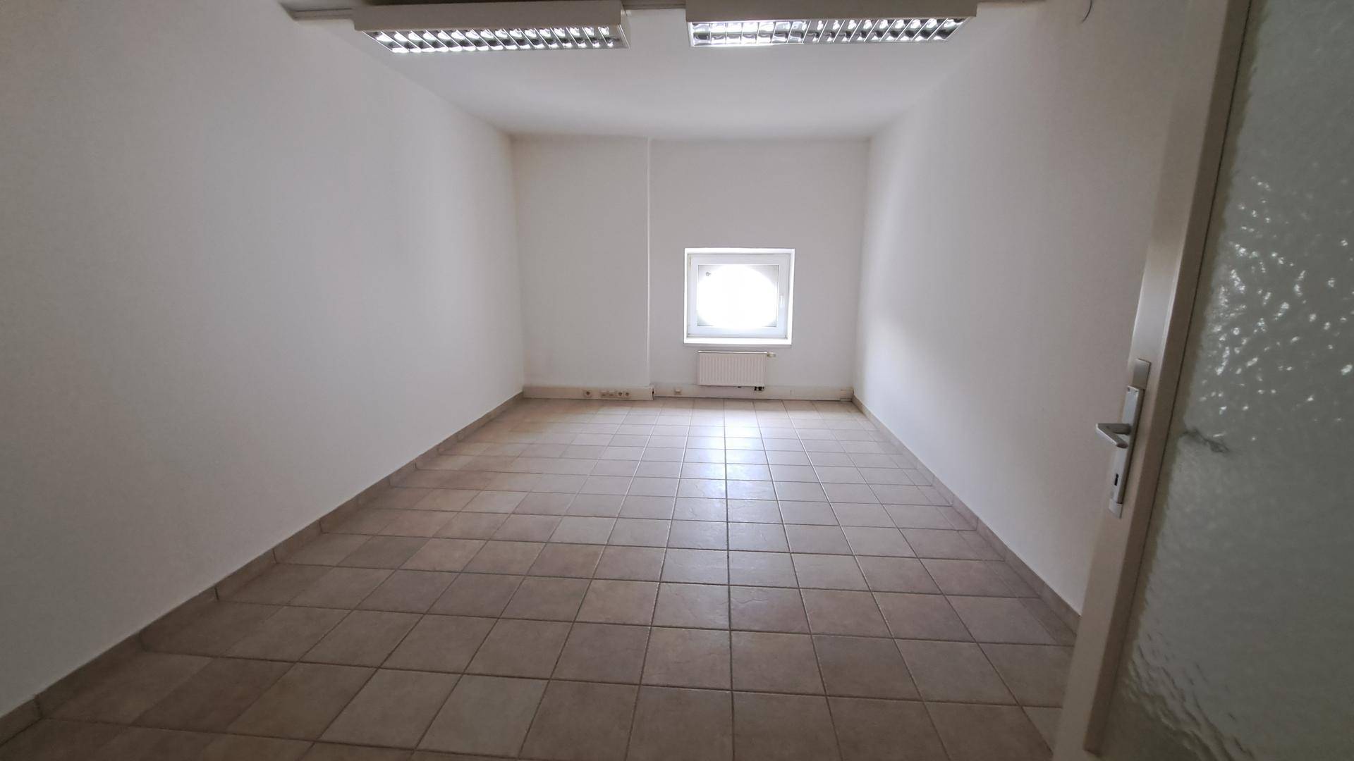 Schmaler Büroraum mit Fliesenboden, weißen Wänden und einem einzelnen runden Fenster.