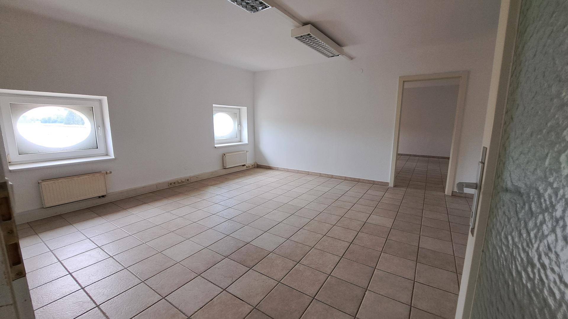 Leerer Büroraum mit Fliesenboden, weißen Wänden und zwei kleinen runden Fenstern.