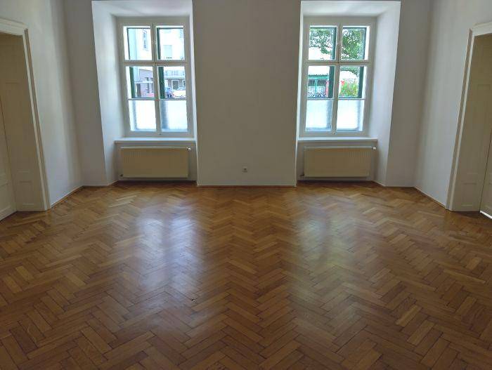 Heller Raum mit Fischgrätparkettboden, zwei Fenstern und weißen Wänden, ideal als Wohnzimmer.