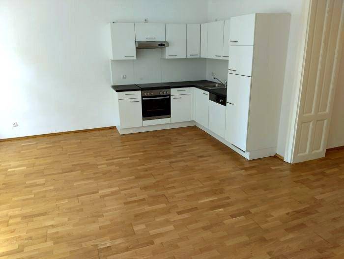 Moderne Einbauküche mit weißen Schränken, dunkler Arbeitsplatte und Parkettboden in einem offenen Raum.
