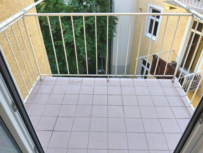 Kleiner Balkon mit Fliesenboden und weißem Geländer, bietet Blick auf den grünen Innenhof.