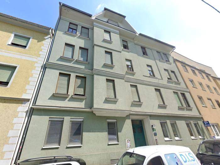 Mehrstöckiges Wohngebäude mit hellgrüner Fassade, vielen Fenstern und Rollläden in einer städtischen Umgebung.
