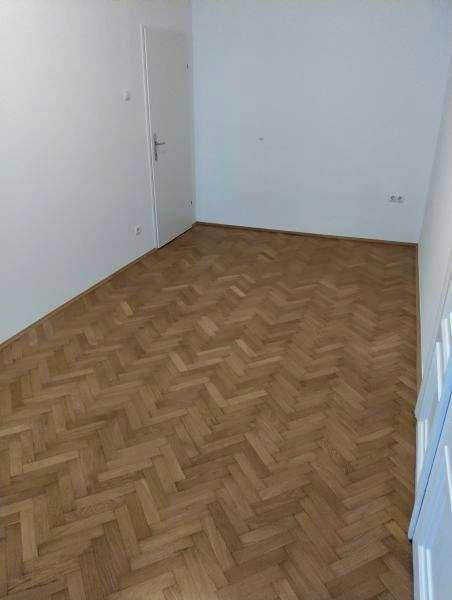 Einfaches Zimmer mit Fischgrätparkett und einer weißen Tür, ideal als Schlafzimmer.