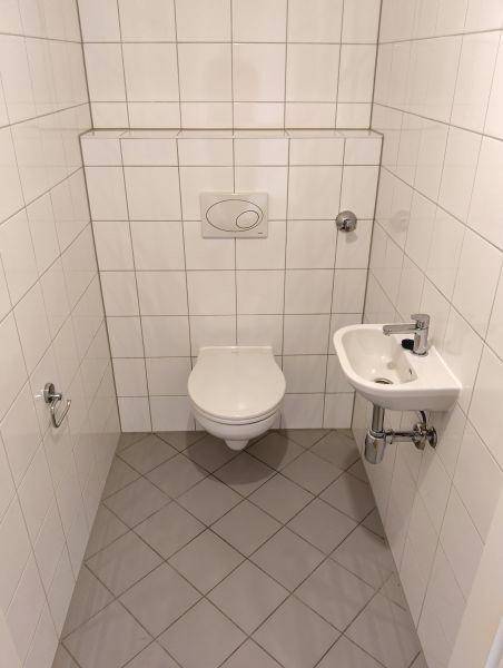 Separates WC mit wandhängender Toilette, kleinem Waschbecken und hellen Fliesen.