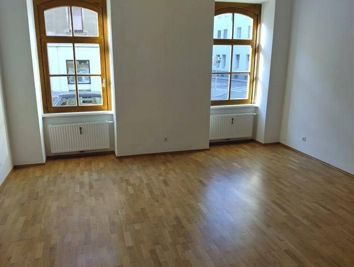 Geräumiger Raum mit zwei großen Fenstern, Parkettboden und Heizkörpern, bietet viel Tageslicht.