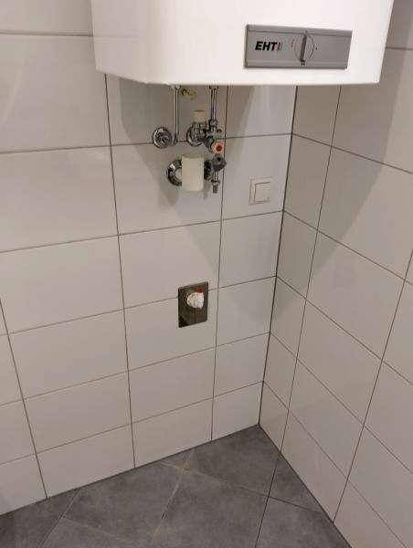 Kleiner Raum mit weißen Wandfliesen, grauen Bodenfliesen und Warmwasserbereiter.