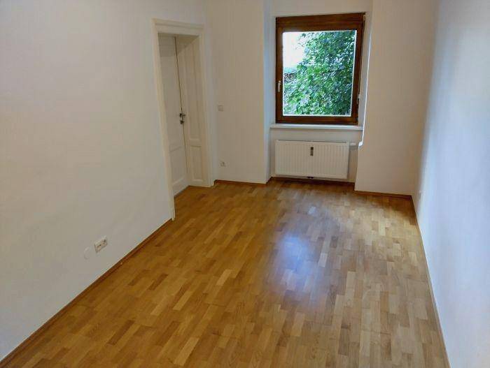 Freundliches Zimmer mit Parkettboden, weißen Wänden und einem Fenster mit grünem Ausblick.
