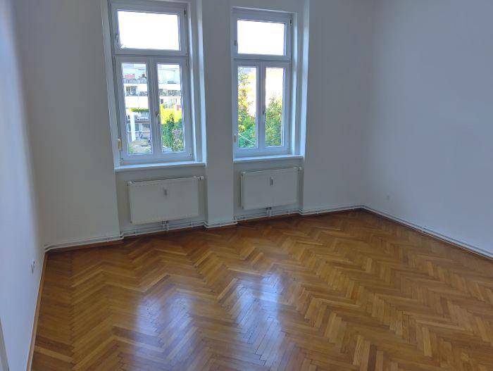 Geräumiges Zimmer mit traditionellem Fischgrätparkett und viel Tageslicht durch die Fenster.