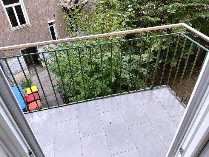 Heller Balkon mit Fliesenboden und grünem Geländer, bietet einen Blick auf den Innenhof und Bäume.