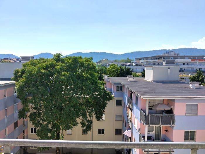 Weitblick über Wohngebäude, grüne Bäume und die umliegende Berglandschaft.