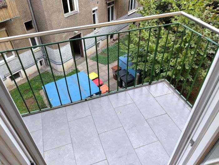 Balkon mit grauen Fliesen und grünem Geländer, bietet einen Blick auf den begrünten Innenhof.