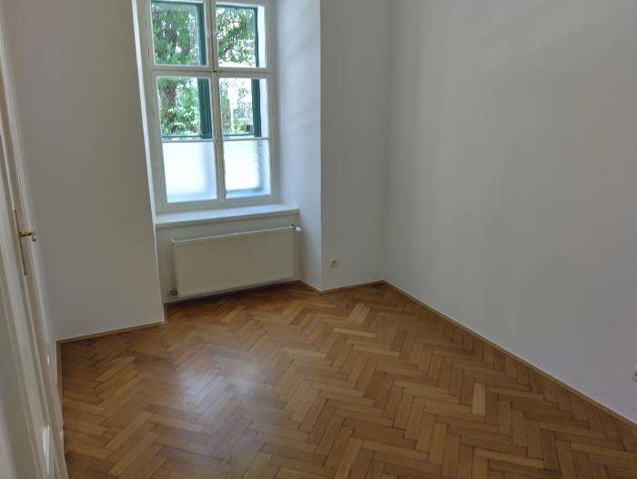 Geräumiger Raum mit Fischgrätparkett, Fenster und Heizkörper, bietet viel Tageslicht.