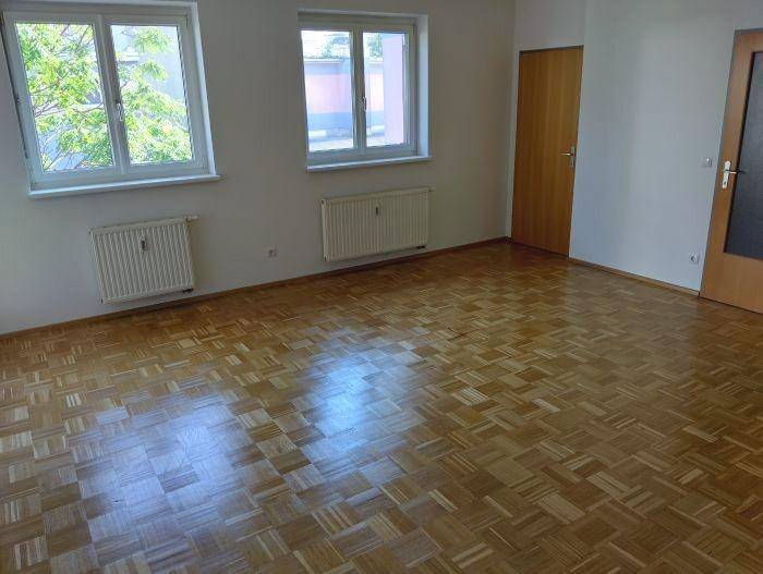 Geräumiges Zimmer mit hellem Parkettboden und zwei großen Fenstern, die viel Licht hereinlassen.
